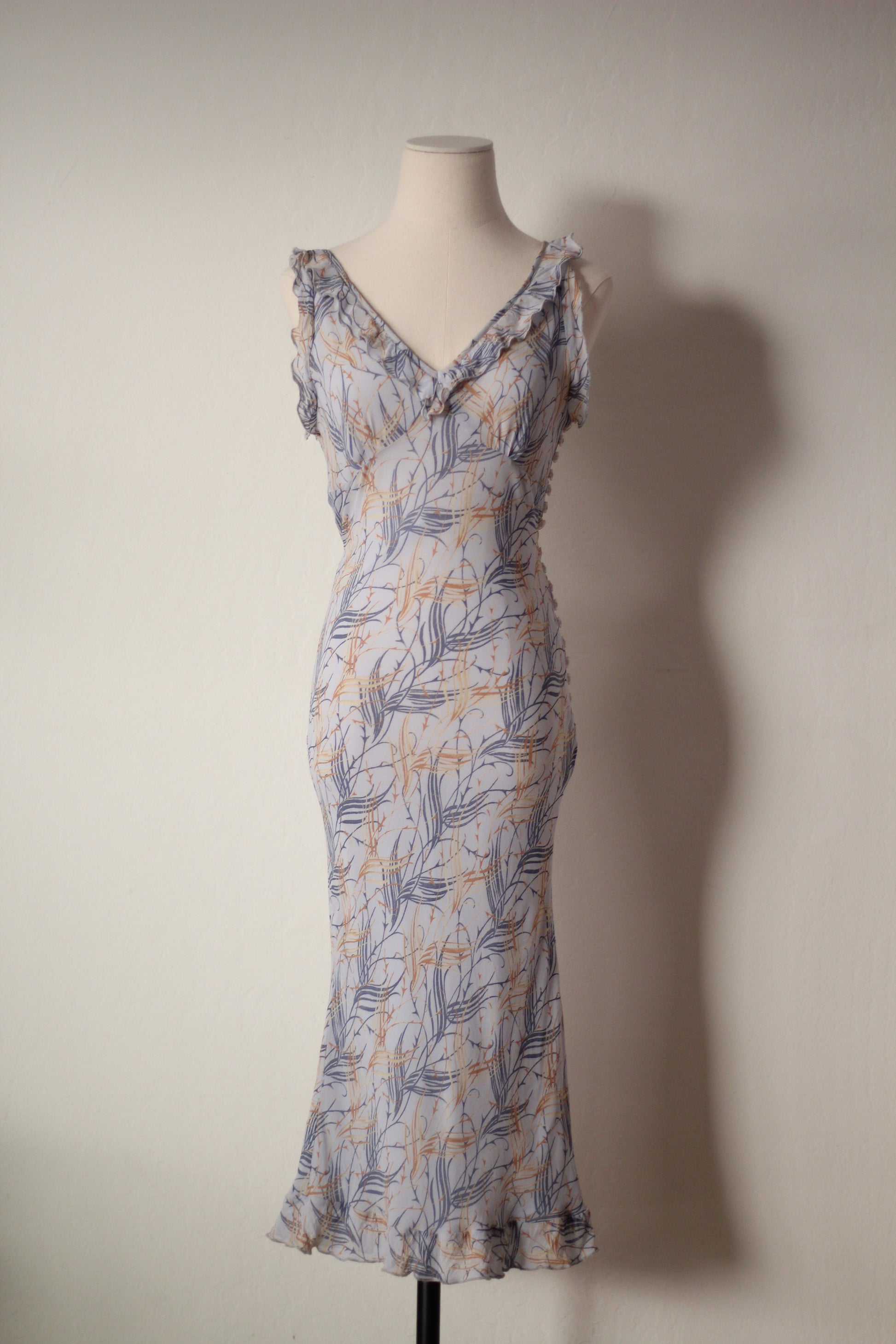 A sleeveless wrap-style maxi dress with a ruffled V-neckline and ruffled straps, featuring a blue-gray and mustard yellow botanical leaf pattern on a light gray base, displayed on a mannequin against a plain wall.