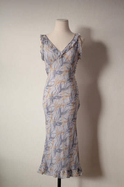 A sleeveless wrap-style maxi dress with a ruffled V-neckline and ruffled straps, featuring a blue-gray and mustard yellow botanical leaf pattern on a light gray base, displayed on a mannequin against a plain wall.