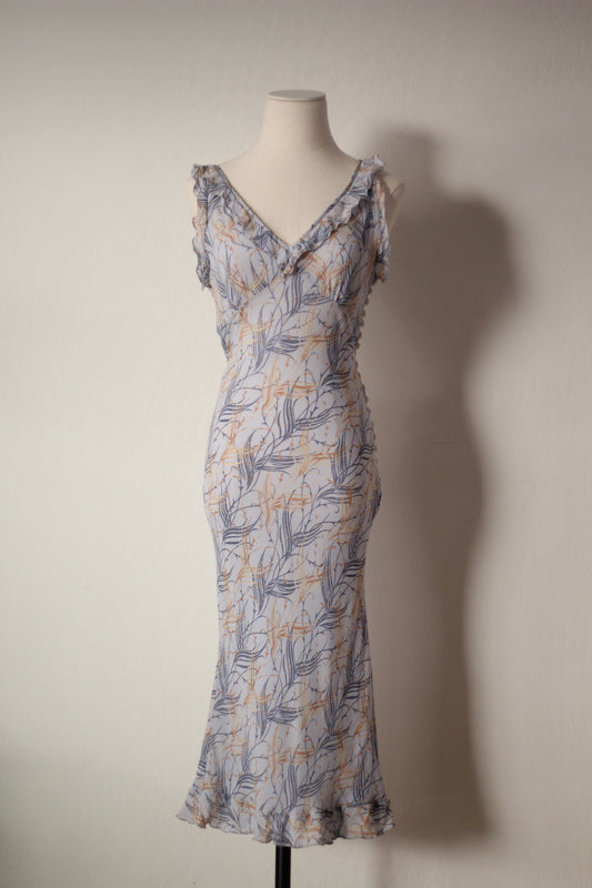 A sleeveless wrap-style maxi dress with a ruffled V-neckline and ruffled straps, featuring a blue-gray and mustard yellow botanical leaf pattern on a light gray base, displayed on a mannequin against a plain wall.