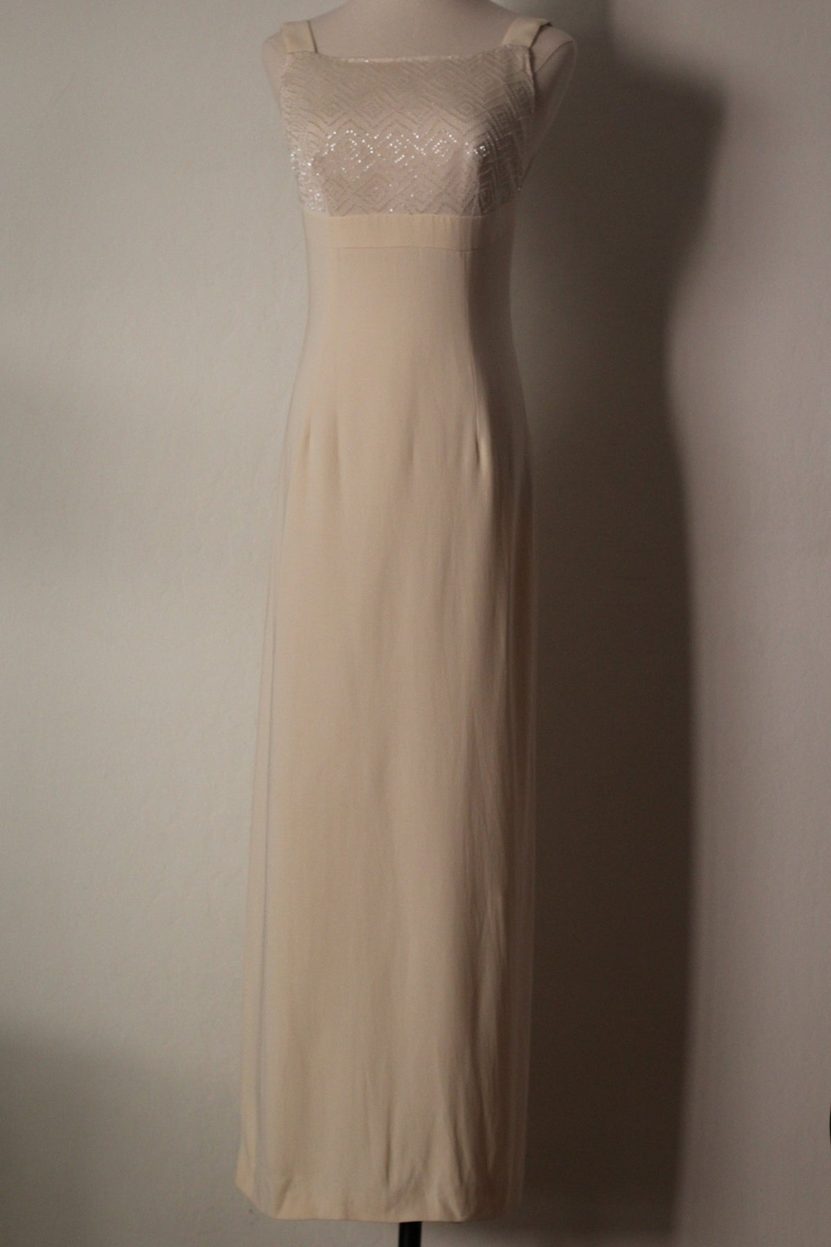 A sleeveless cream-colored maxi dress with a sheer lace bodice and a solid satin-like skirt, displayed on a mannequin against a plain wall.