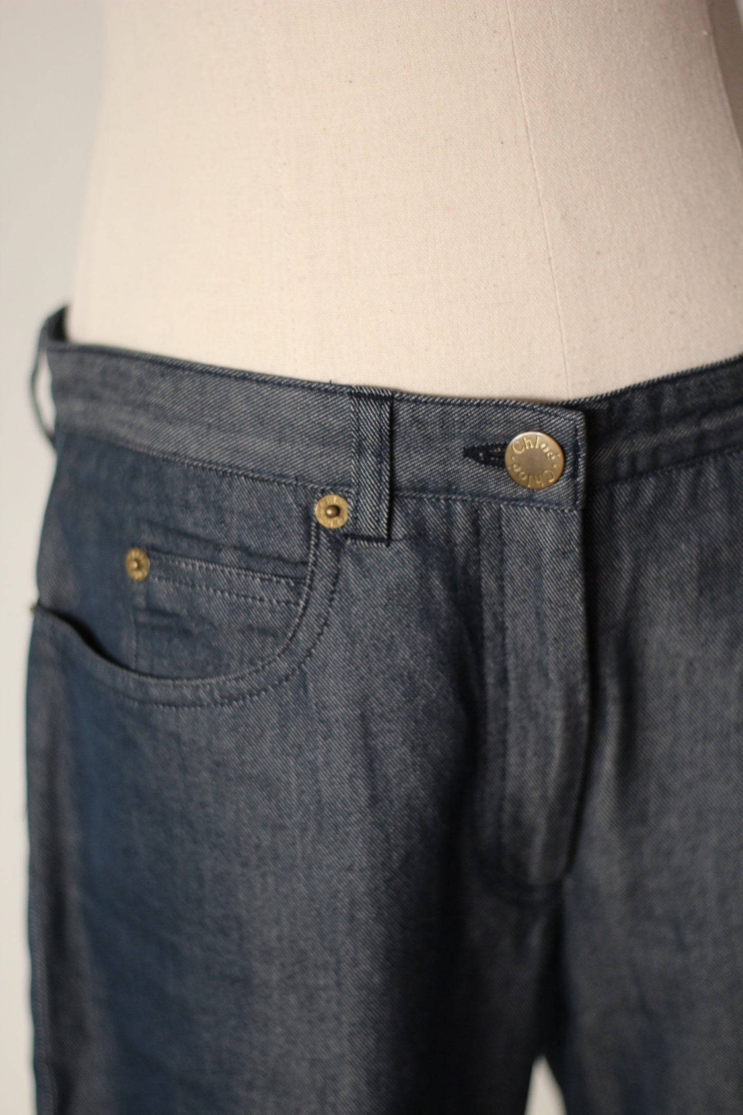 Close-up of dark blue denim jeans on a cream dress form, showing the waistband, belt loop, button, front pocket, and rivets.