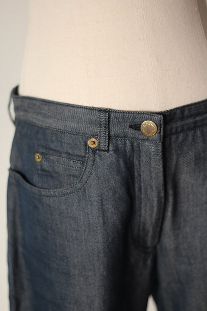 Close-up of dark blue denim jeans on a cream dress form, showing the waistband, belt loop, button, front pocket, and rivets.