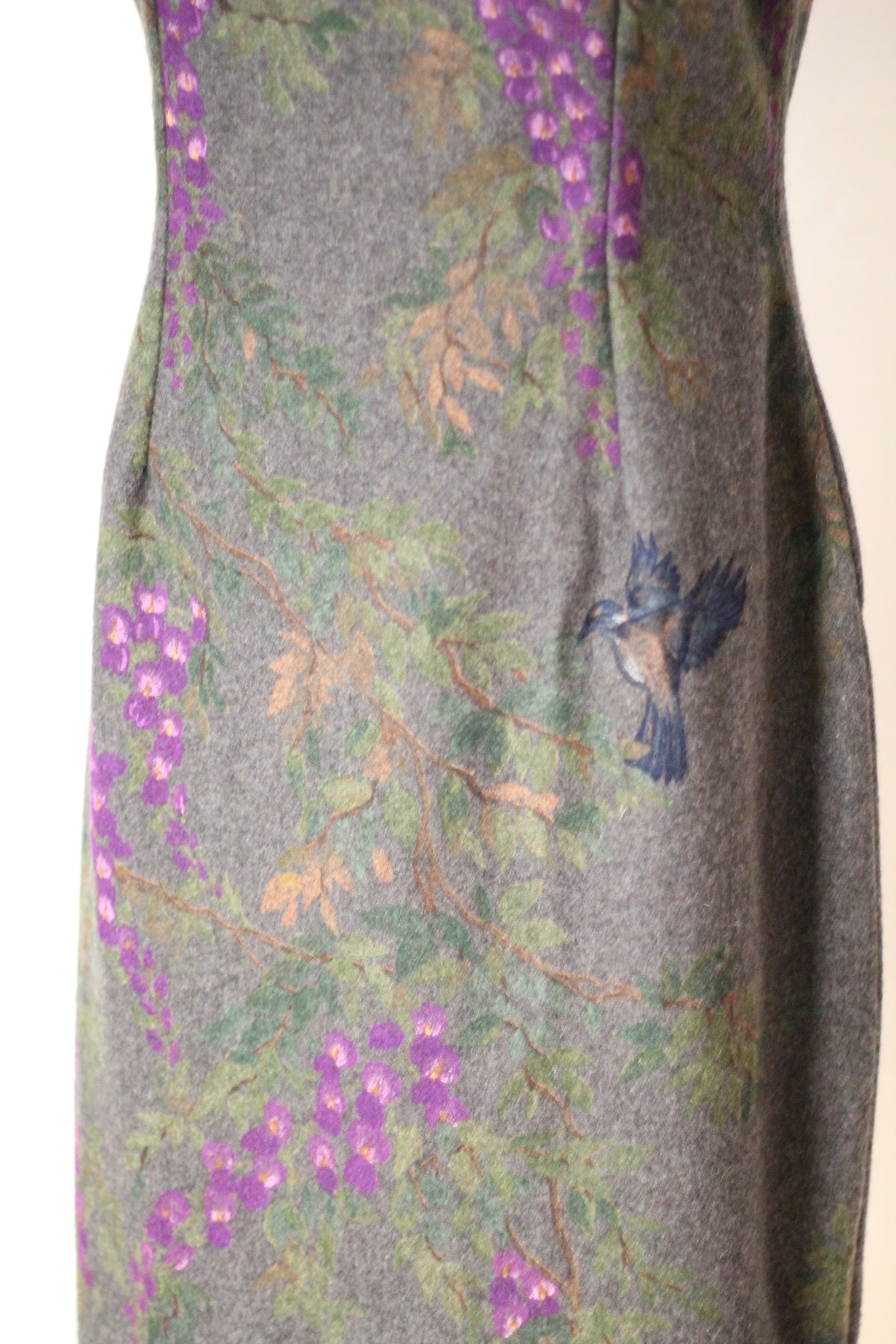 Close-up of vintage Dolce & Gabbana Fall/Winter 1998 runway dress showing a painted blue bird surrounded by purple wisteria flowers, green foliage, and branches on gray wool fabric.