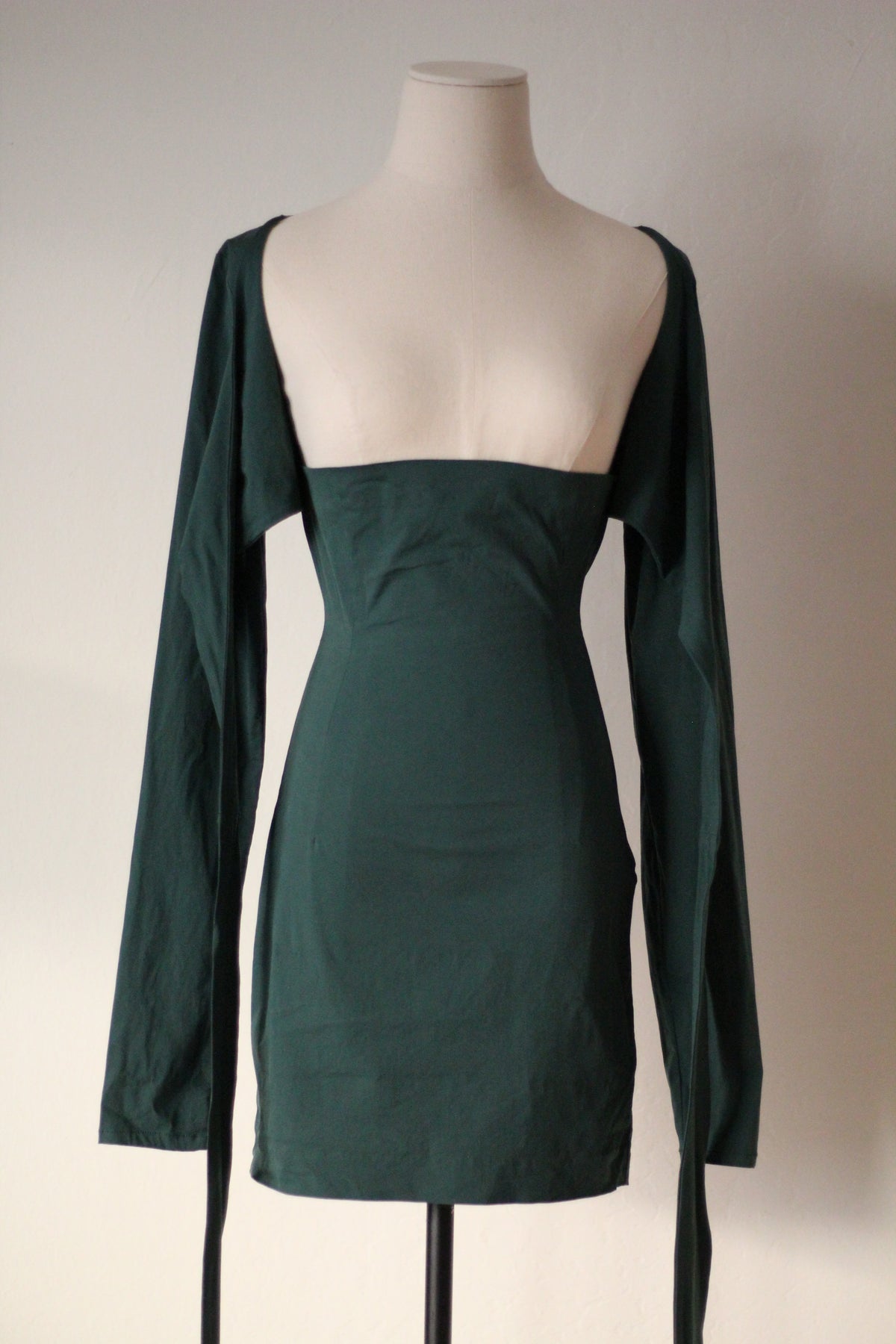 Dark green long-sleeve mini dress with a square neckline, fitted bodice, and draped side ties, displayed on a mannequin